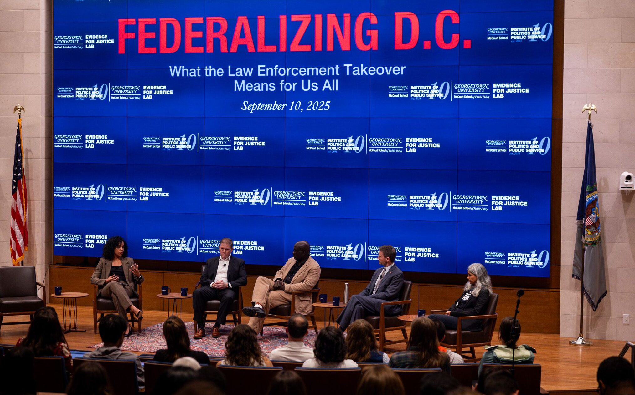 A wide shot of a panel discussion held on a stage with large blue digital screens in the background displaying the Evidence for Justice Lab and Institute of Politics and Public Service logos. From left to right: Dr. Andrea M. Headley (moderator) is speaking; seated next to her are Councilmember Charles Allen, Jamelle Bouie, Steven Sund, and Keya Chatterjee.